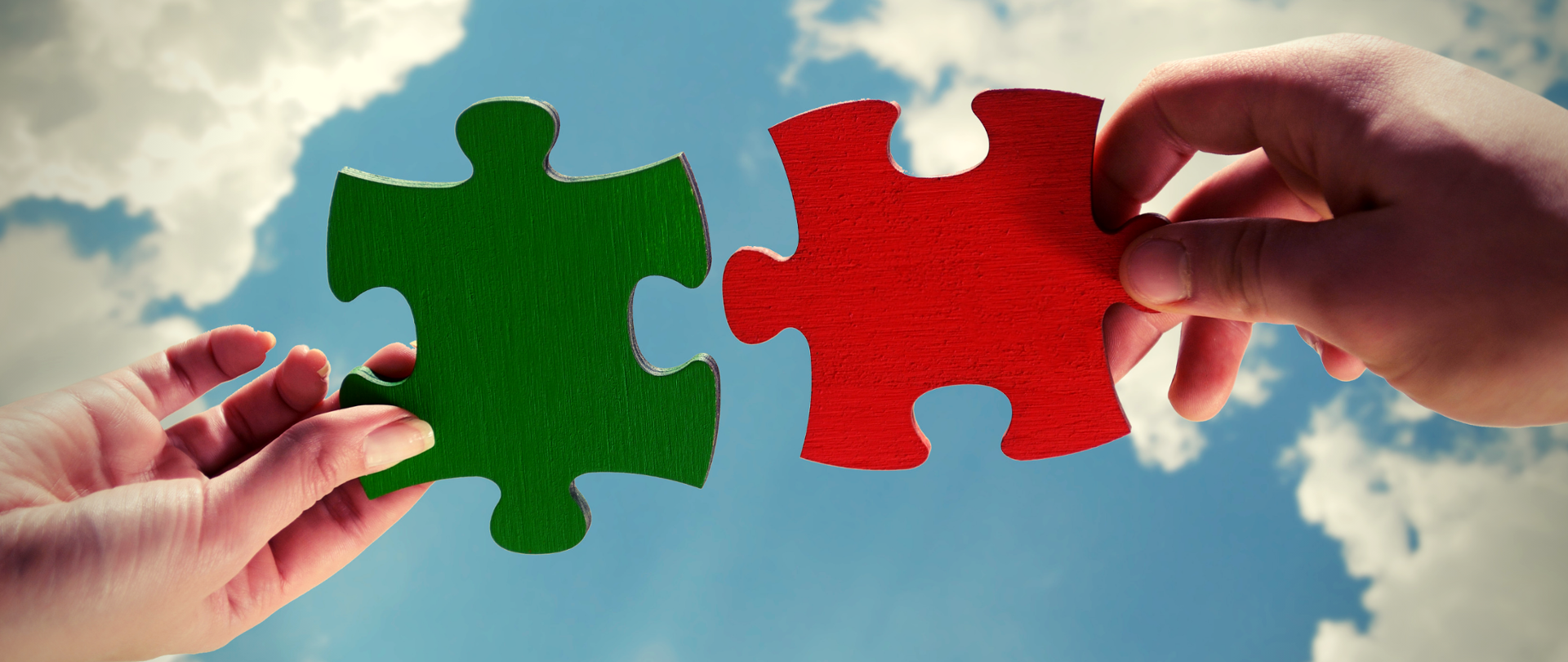 Dwie dłonie na tle nieba. Każda trzyma puzzel, jeden jest w kolorze zielonym, drugi czerwonym