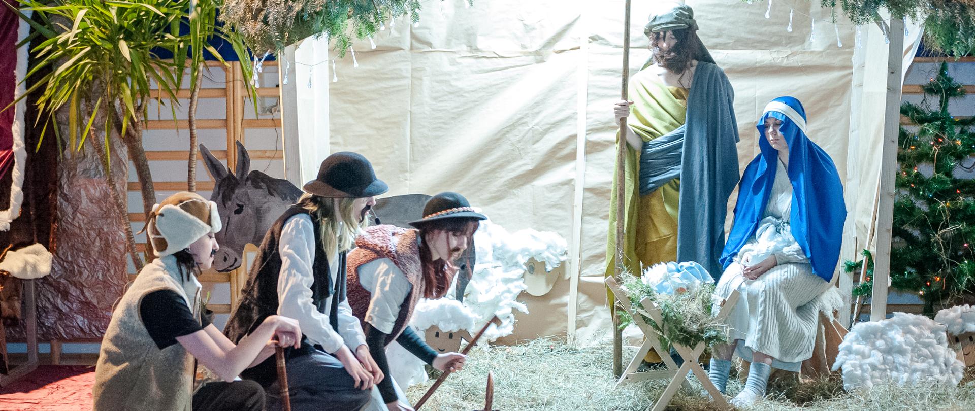 Na tle stajenki widać Maryję i Józefa oraz żłobek z Dzieciątkiem. Po lewej stronie klęczą trzej pasterze. Widać również przyniesione przez nich dary np. kosz i ser. 