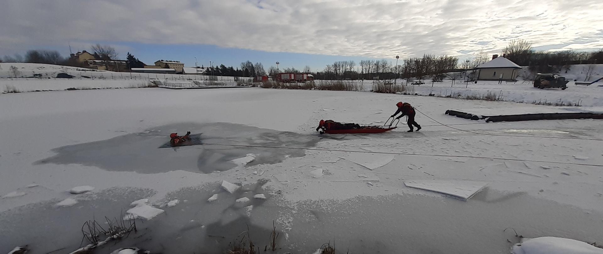 Na zdjęciu sceneria zimowa, zamarznięty akwen wodny, w przerębli w wodzie strażak w stroju ochronnym, do niego na saniach zbliża się dwóch strażaków. 