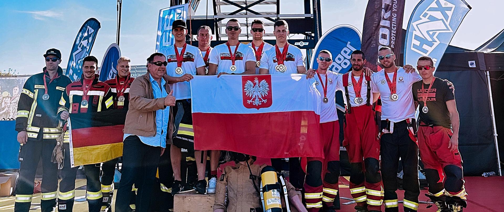 Podium na którym widoczne są reprezentacje strażaków z Polski i Niemiec. Polskie drużyny stoją na najwyższym i drugim miejscu podium, trzecie miejsce zajmuje drużyna niemiecka. Reprezentacje drużyn prezentują przed sobą flagi narodowe. W tle widoczna jest wspinalnia oraz banery reklamowe sponsorów. 