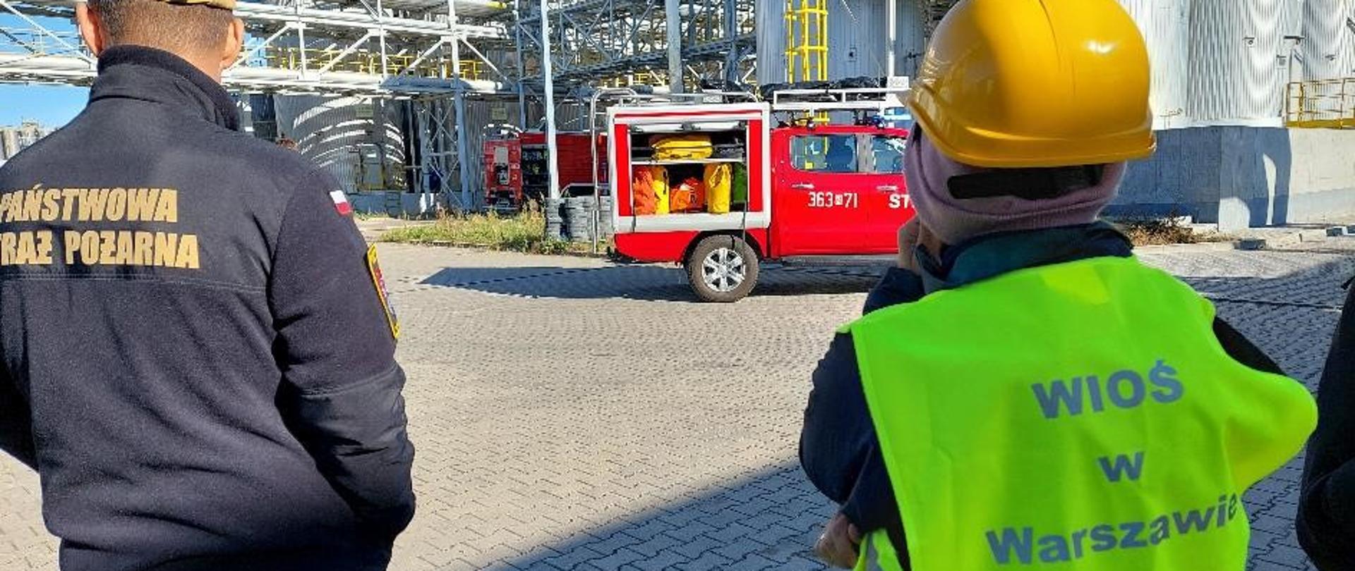 Inspektor Wojewódzkiego Inspektoratu Ochrony Środowiska w Warszawie oraz funkcjonariusz Państwowej Straży Pożarnej monitorują działania prowadzone przez Państwową Straż Pożarną. W tle widać wozy bojowe PSP oraz stalowe, srebrne silosy o kilkunastometrowej wysokości wraz z instalacjami je łączącymi.