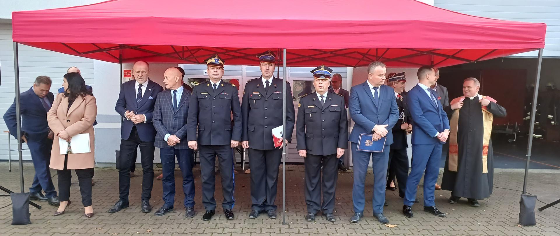 grupa ludzi stoi po namiotem , wśród grupy znajdują się strażacy , ksiadz oraz osoby cywilne, w tle podniesiona brama garażowa remizy