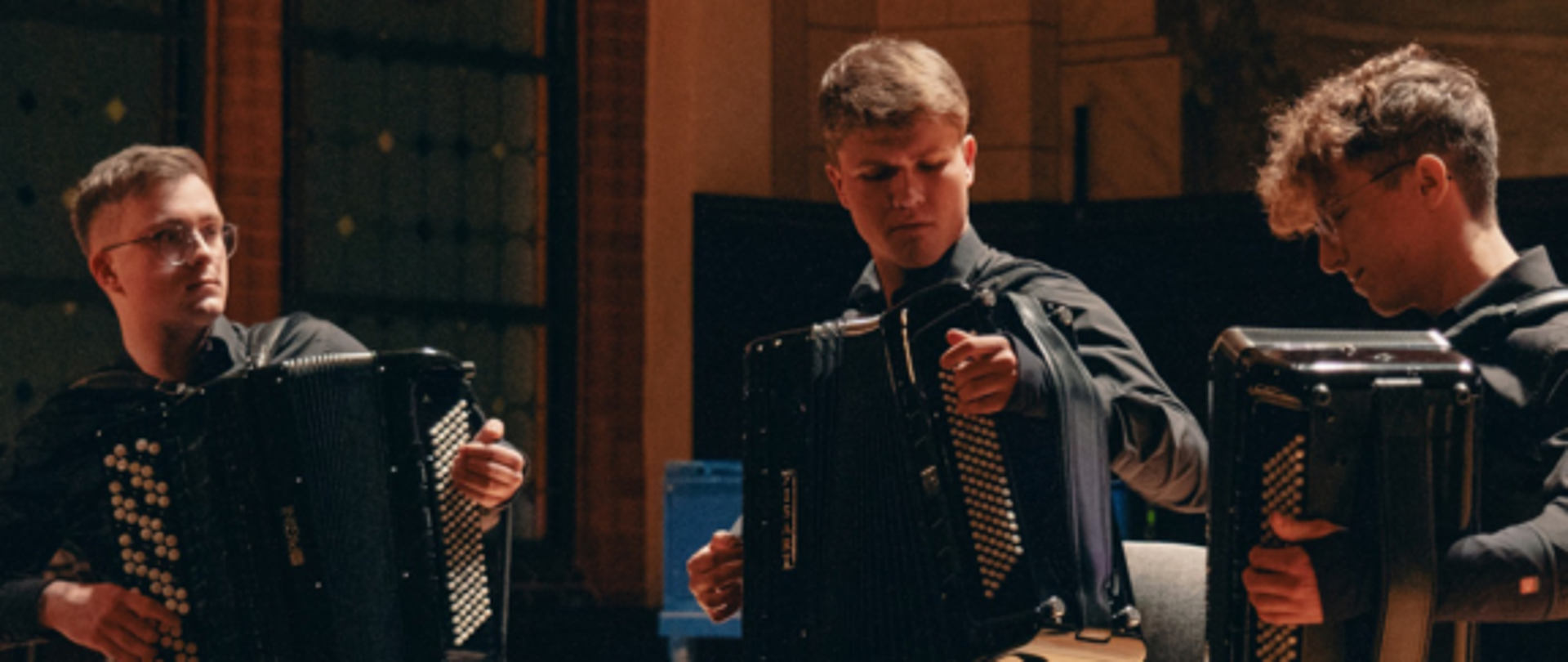 u góry na czerwonym tle białe napisy, pośrodku zdjęcie koncertującego tria akordeonowego, na dole na białym tle czerwone napisy