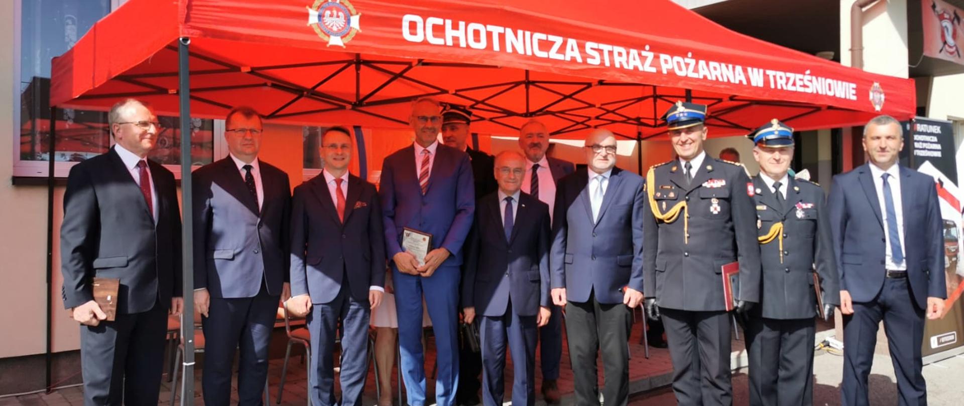 Zdjęcie zrobione na zewnątrz. Pod namiotem z napisem Ochotnicza Straż Pożarna w Trześniowie stoją uczestnicy uroczystości. 