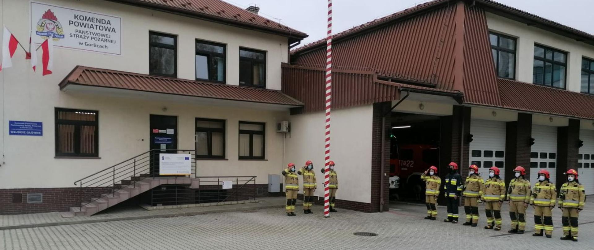 Na zdjęciu widoczni strażacy na tle budynku komendy. Na środku zdjęcia masz z flagą i salutujący trzej strażacy. Po prawej stronie zmiana służbowa w jednym szeregu z salutującym dowódcą zmiany służbowej.