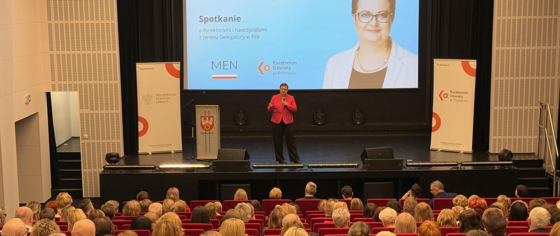 Widok na scenę na której przemawia wiceminister Lubnauer. W tle duży ekran ze zdjęciem Katarzyny Lubnauer i tytułem spotkania. Po lewej stronie stoi mównica z herbem powiatu i plansza z napisem Wielkopolski Kurator Oświaty