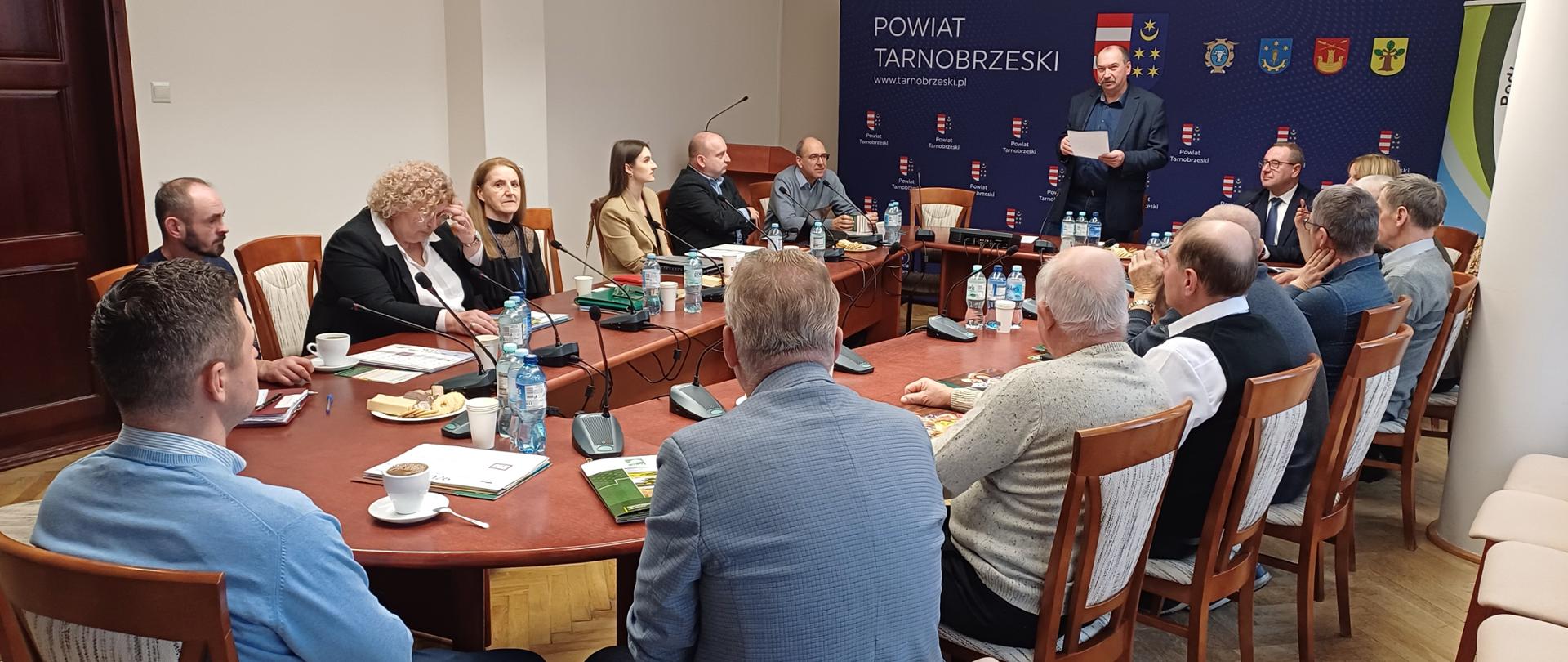 Grupa ludzi siedząca przy stołach. Znajdują się w jasnym pomieszczeniu. W tle stoi duży granatowy baner a przed nim stoi mężczyzna. Na stołach stoją mikrofony oraz wody w butelkach.