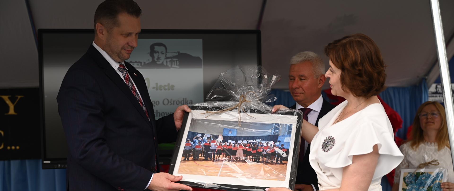 Minister Czarnek stoi naprzeciwko kobiety w białej sukience, razem trzymają kolorowy obrazek.