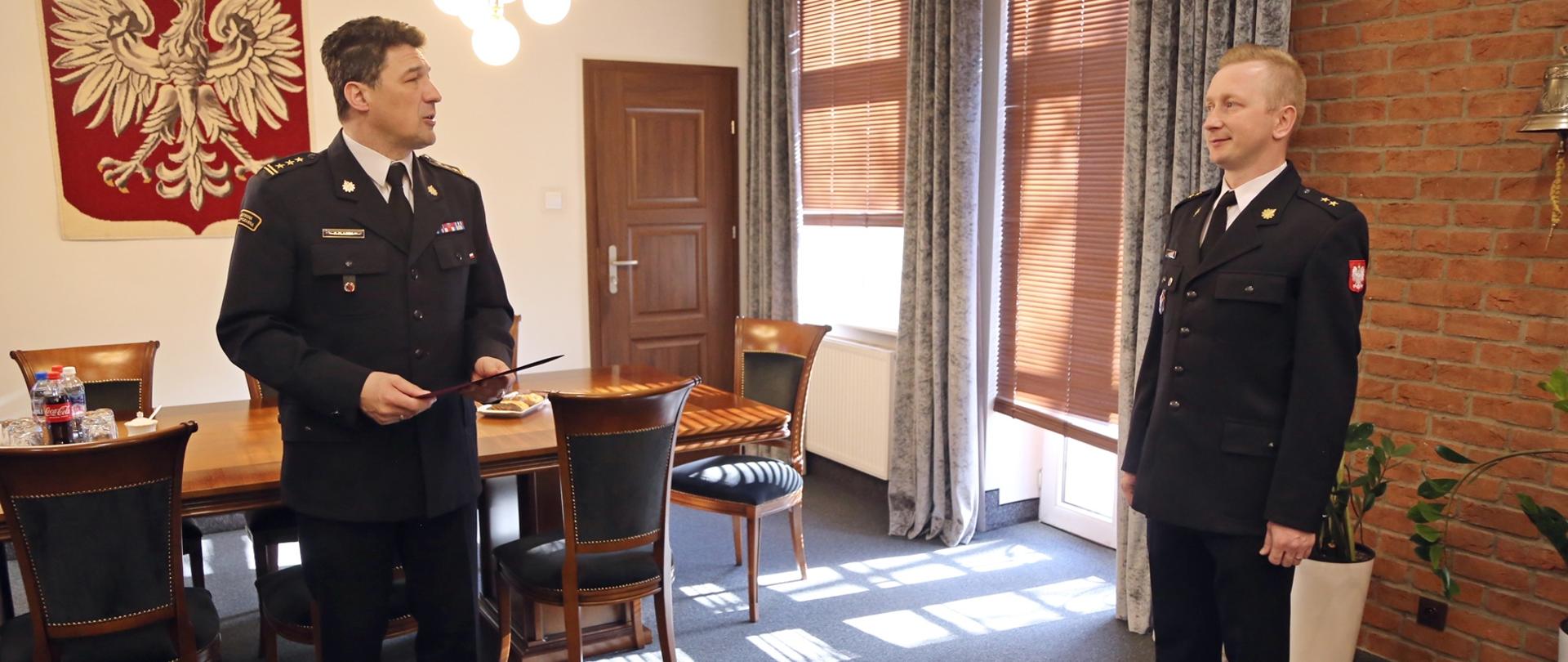 W gabinecie Komendanta Centralnej Szkoły PSP. Komendant stojący po lewej stronie zwraca się do nagradzanego funkcjonariusza stojącego z prawej strony