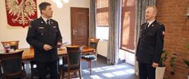 W gabinecie Komendanta Centralnej Szkoły PSP. Komendant stojący po lewej stronie zwraca się do nagradzanego funkcjonariusza stojącego z prawej strony