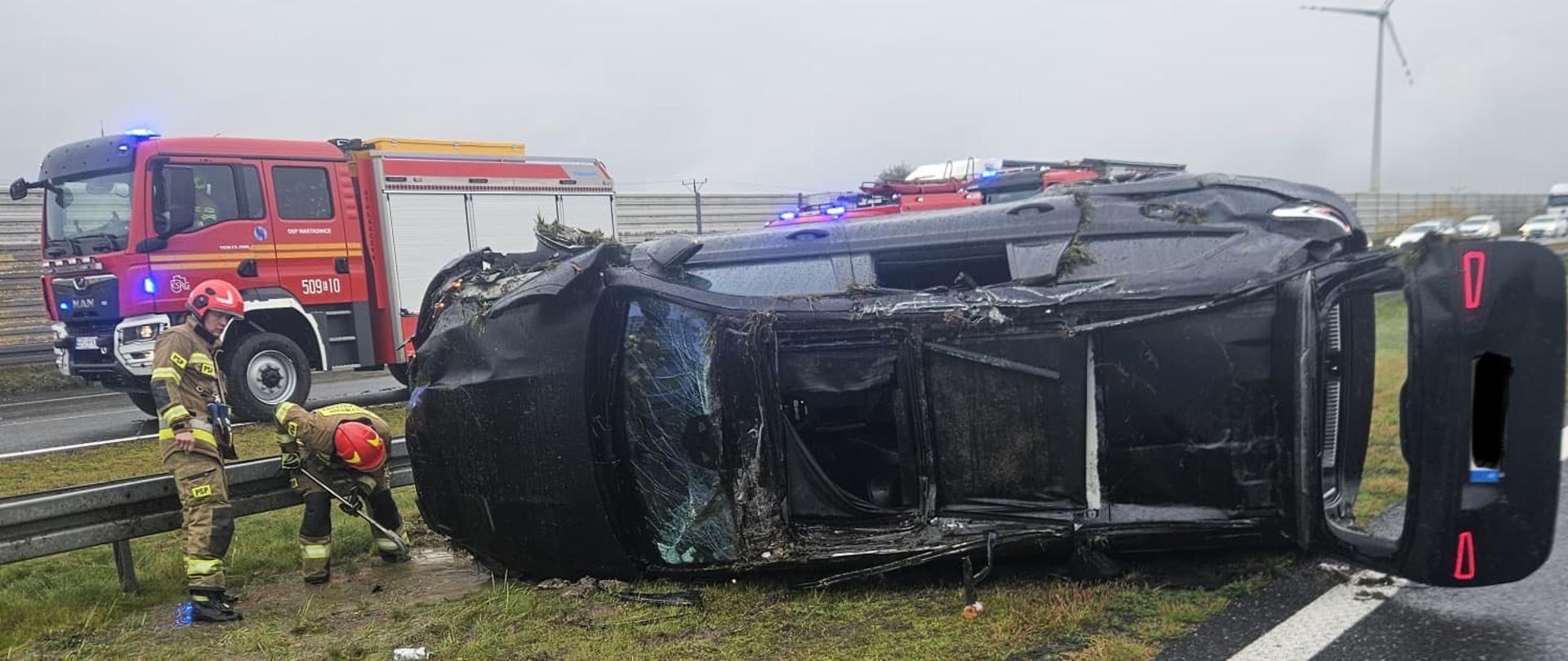 Czarny samochód leży na boku przednia szyba jest popękana a dach powgniatany . z przodu samochody stoją dwaj strażacy w tle widać dwa wozy strażacki 