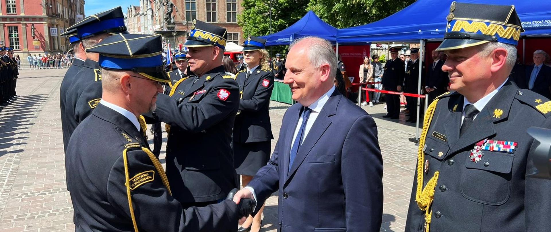 Zastępca komendanta głównego PSP wraz z wojewodą i pomorskim komendantem wojewódzkim PSP wręcza odznaczenie strażakowi PSP, w tle inni strażacy oraz uczestnicy obchodów
