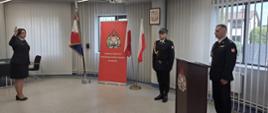 Zdjęcie zrobione w pomieszczeniu strażnicy na tle godła Rzeczpospolitej Polskiej, dwóch Flag Rzeczpospolitej Polskiej, po lewej stronie banneru Komendy Powiatowej PSP w Lublińcu. Po lewej stronie nowoprzyjęty strażak stojący na baczność i ubrany w umundurowanie galowe składa ślubowanie podnosząc prawą rękę, po prawej stronie Komendant w ubraniu galowym stojący za mównicą oraz dowódca uroczystości w umundurowaniu galowym z pasem i w czapce.