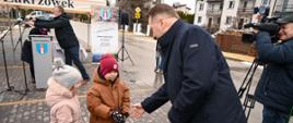 Zdjęcie na dworze, minister Czarnek nachyla się do małego dziecka w brązowej kurtce trzymającego bukiecik kwiatów, obok filmujący go mężczyzna z kamerą, w tle budynki.