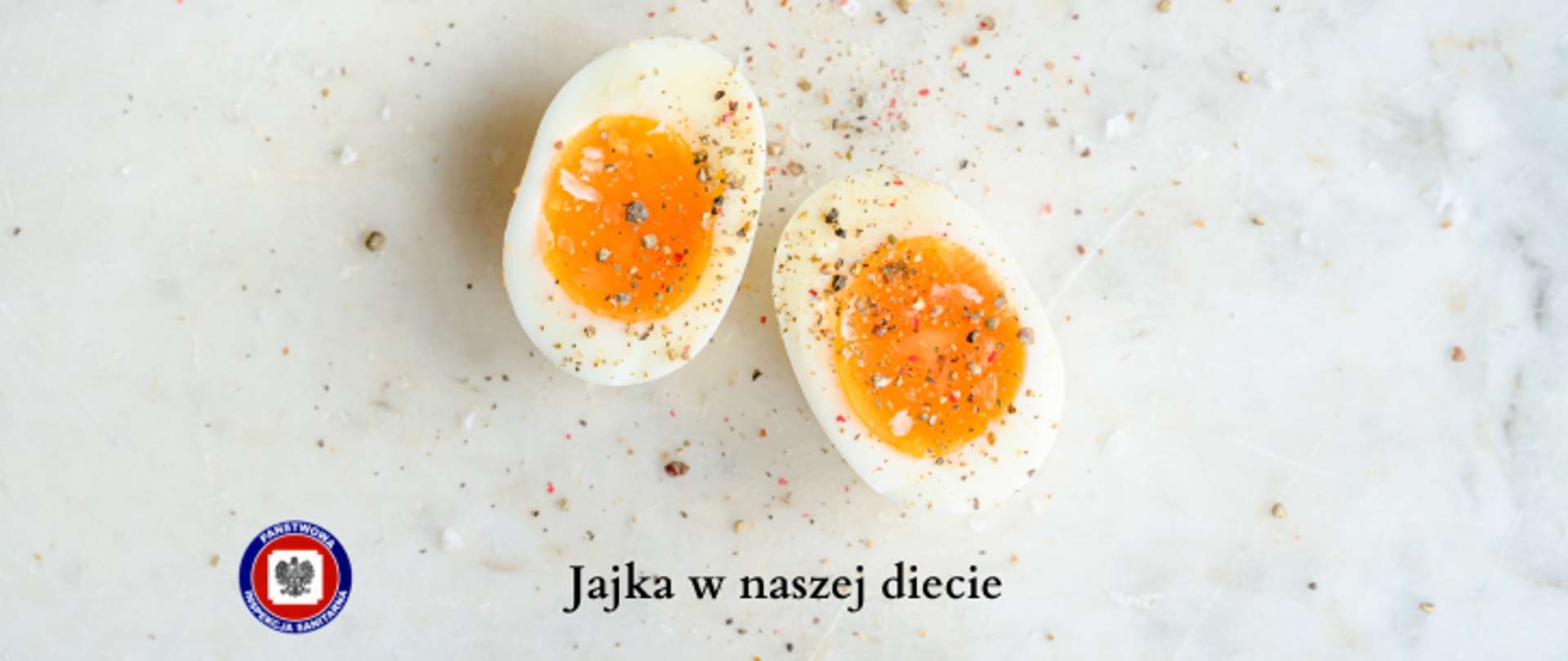 Na jasnym marmurowym stole leży rozkrojone jajko posypane przyprawami, poniżej ciemny napis Jajka w naszej diecie, z jego lewej logo Państwowej Inspekcji Sanitarnej.
