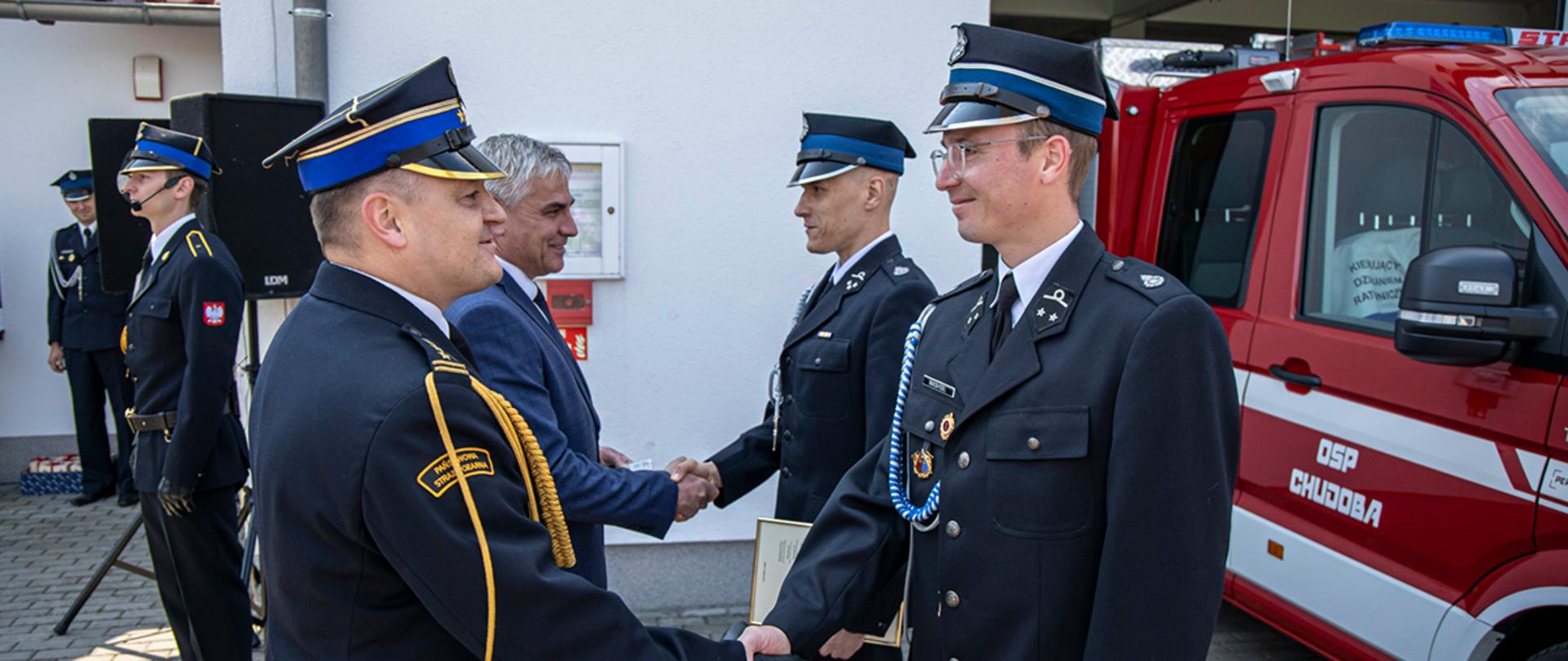 Opolski Komendant Wojewódzki PSP w Opolu st. bryg. Krzysztof Kędryk oraz Wójt Gminy ściskają dłonie druhów OSP (w mundurach wyjściowych) stojących przy samochodzie ratowniczo-gaśniczym z napisem OSP Chudoba wystawionym do połowy przed garaż jednostki. W tle budynek remizy OSP oraz dowódca uroczystości. 
