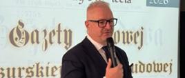 Mężczyzna w granatowej marynarce, wojewoda, przemawia przez mikrofon na tle prezentacji dotyczącej wydarzenia 