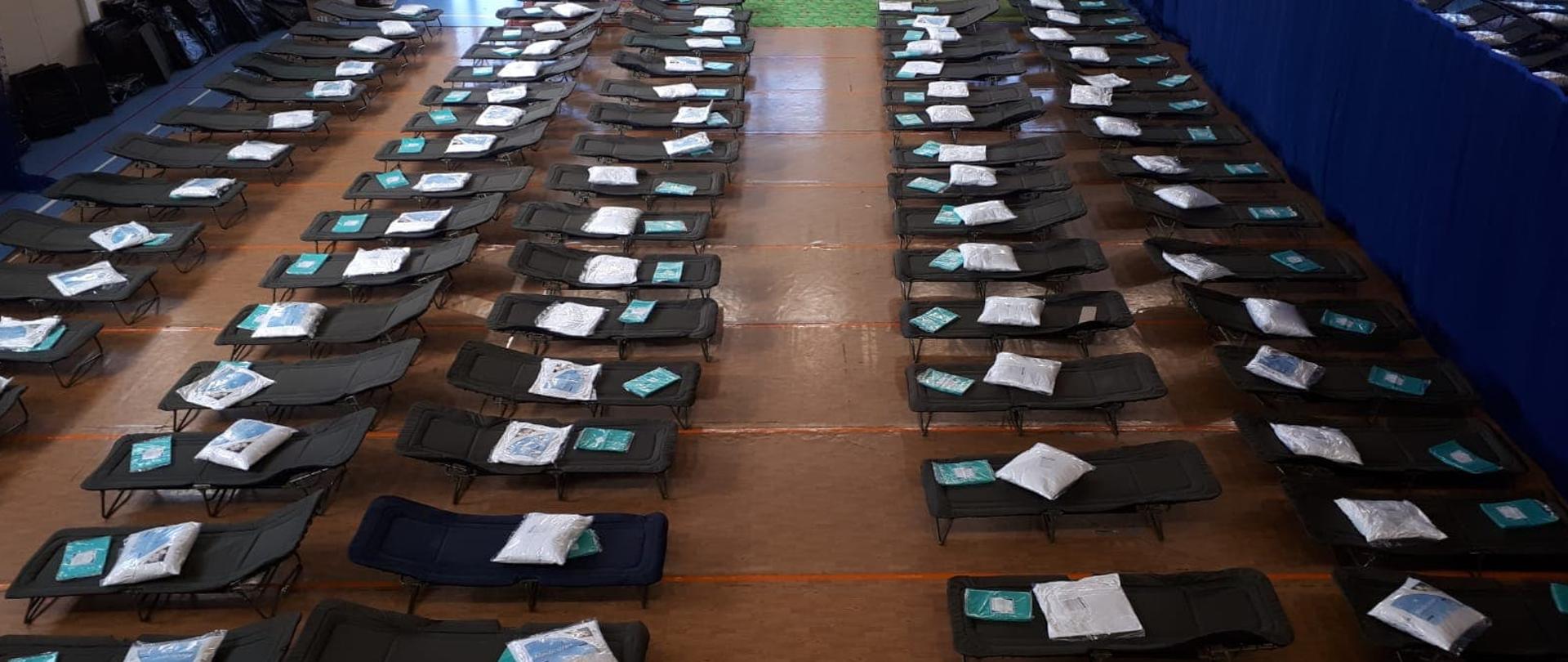 Na zdjęciu widoczne są łóżka ustawione obok siebie na hali. 