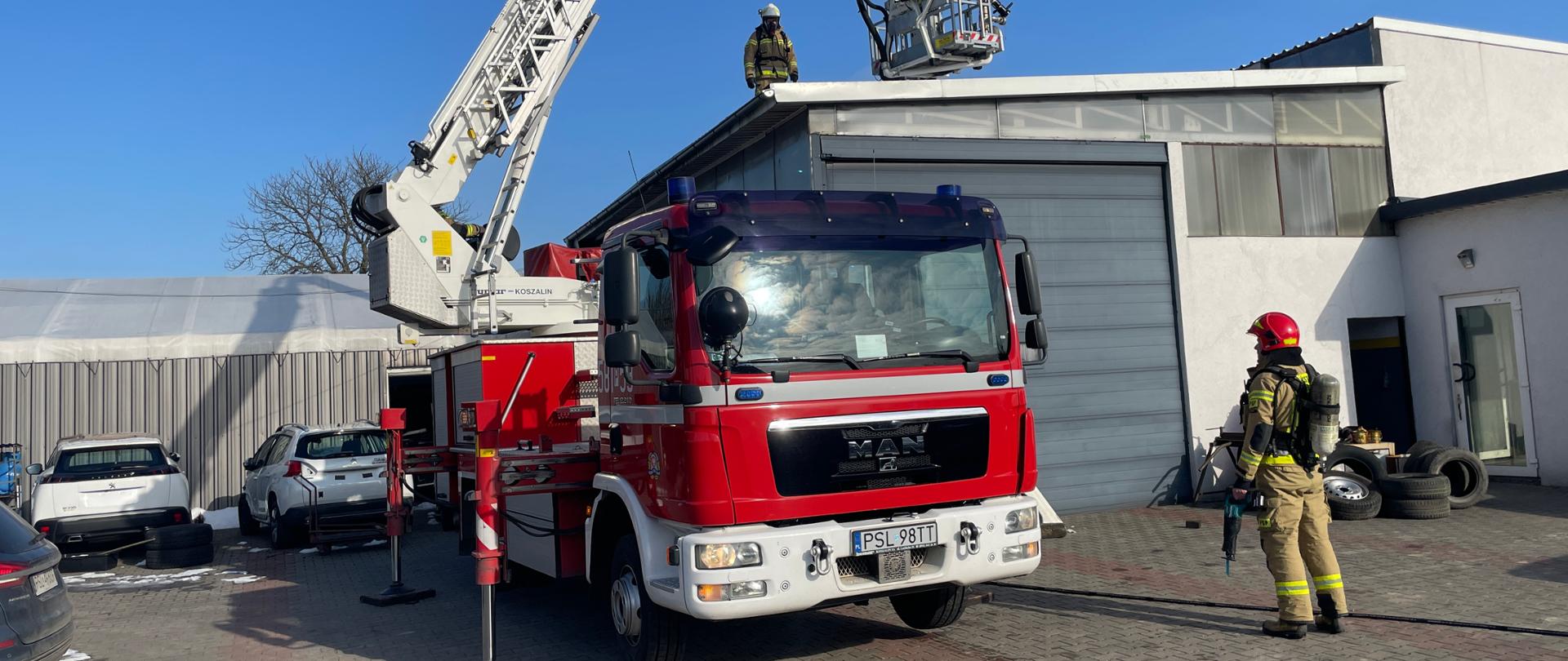 samochód strażacki podnośnik stoi na placu przy budynku na dachu jest jeden strażak drugi stoi przy samochodzie za podnośnikiem samochody po prawej stronie wejście do budynku gdzie lezy kilka opon