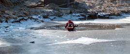 Na zdjęciu widać strażaków w skafandrze suchym na akwenie wodnym który pokryty jest lodem. Jeden ze strażaków porusza się na saniach lodowych, drugi znajduje się w przeręblu.