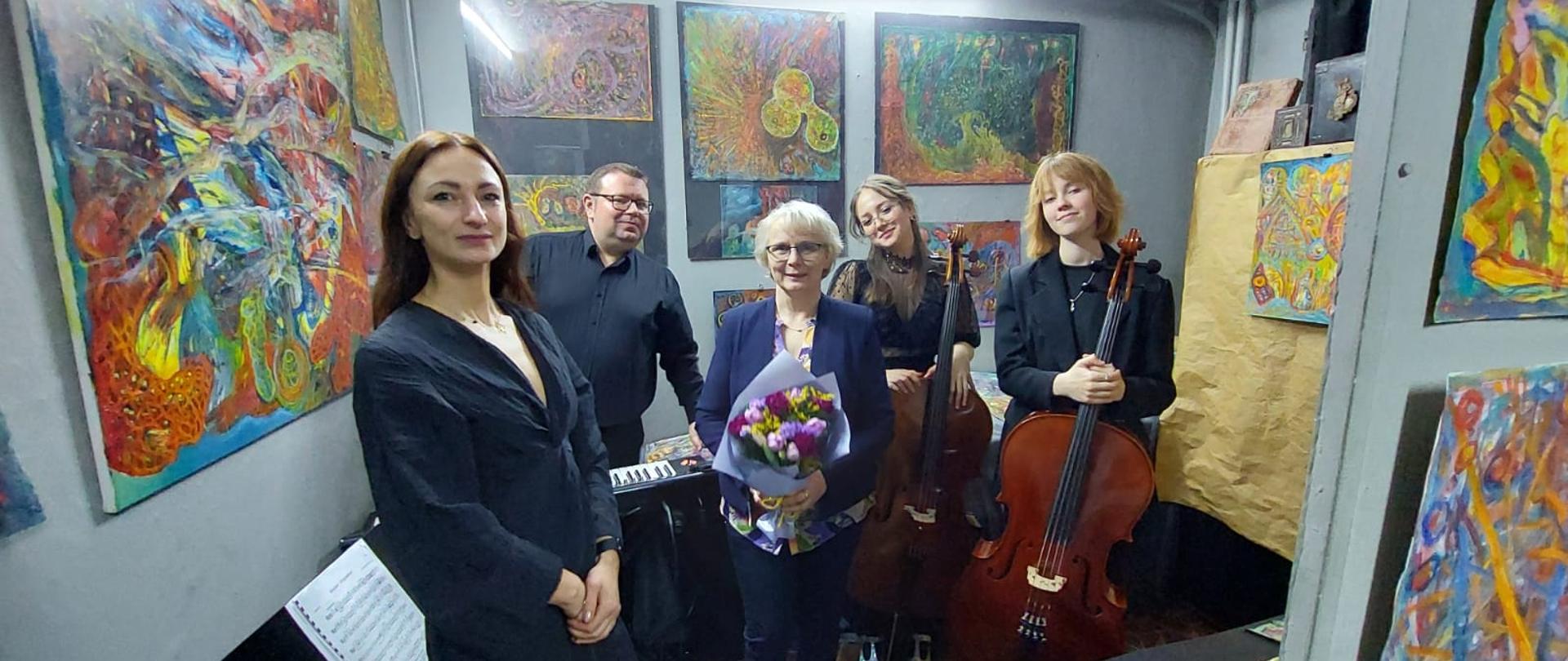 Dwie kobiety, z których jedna trzyma bukiet kwiatów, mężczyzna i dwie nastolatki trzymające wiolonczele stoją w pomieszczeniu, na którego ścianach wiszą kolorowe obrazy.