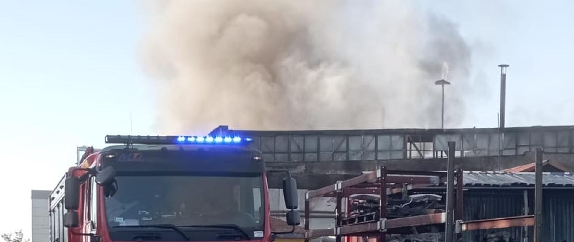 Zdjęcie przedstawia teren zakładu gdzie doszło do pożaru. W głębi hala nad którą unosi się gęsty dym. Przed halą stoi samochód pożarniczy a przy nim strażak.