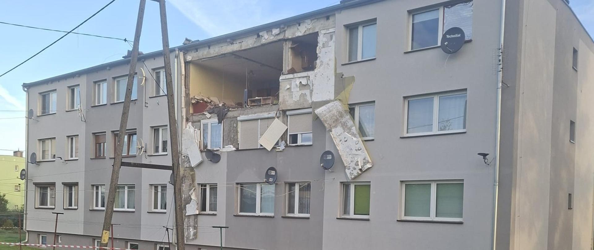 Trzypiętrowy, szary budynek mieszkalny z widocznymi poważnymi uszkodzeniami elewacji na najwyższym piętrze. W ścianie znajduje się duża wyrwa odsłaniająca wnętrze mieszkania, a wokół niej wiszą uszkodzone płaty elewacji i rolety. Przed budynkiem stoi drewniany słup telegraficzny, a na trawniku oraz wokół obiektu rozrzucone są gruzy i fragmenty elewacji. Obszar przed budynkiem został wygrodzony biało-czerwoną taśmą ostrzegawczą.
