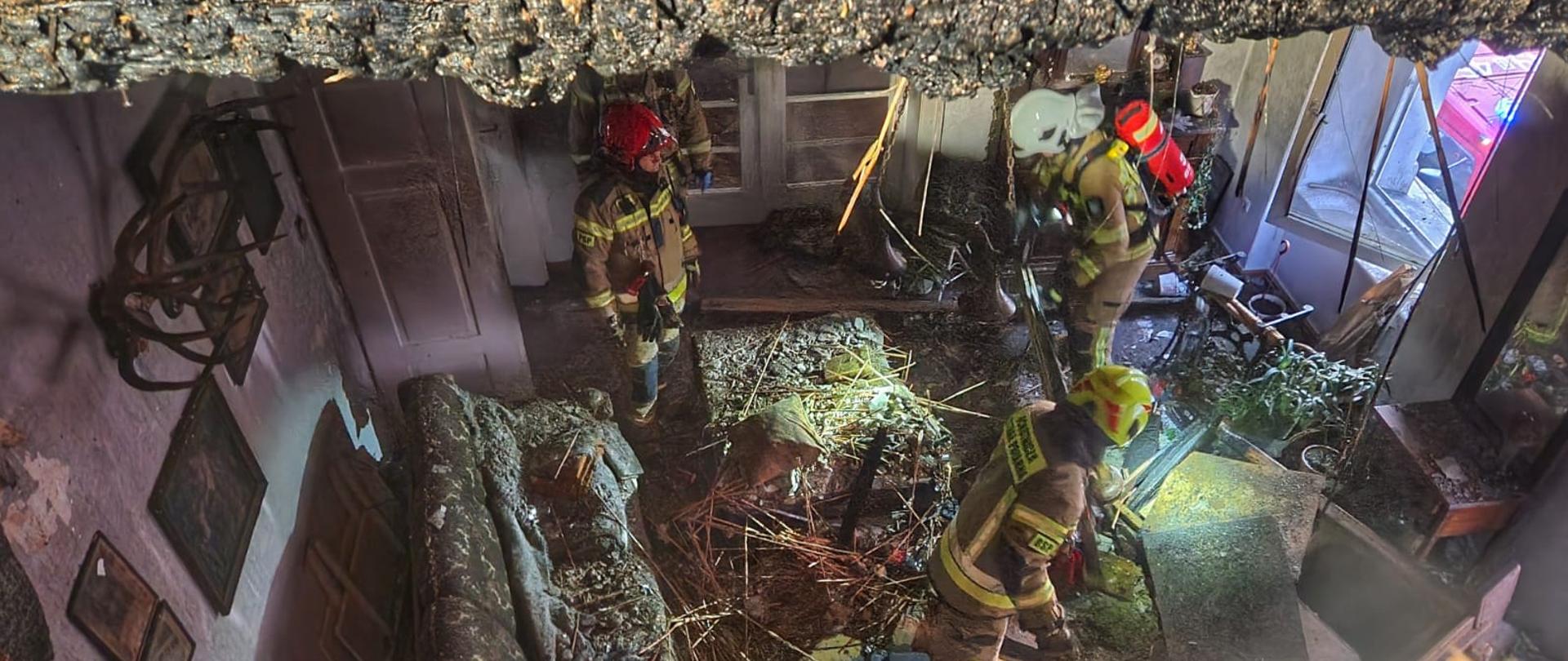 Obraz przedstawia mieszkanie po pożarze. Wewnątrz strażacy w umundurowaniu specjalnym. Zdjęcie z mieszkania powyżej zrobione przez dziurę w stropie.