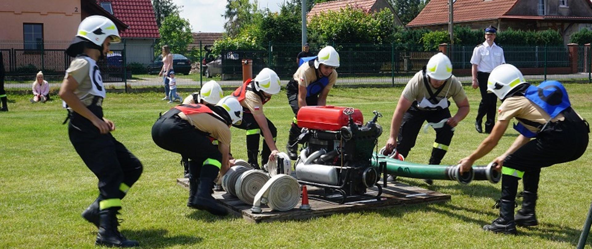 Na trawie ustawiony podest na którym znajduje się sprzęt przeznaczony do wykonania ćwiczenia bojowego, motopompa, 2 węże ssawne 2 odcinki W-75, dwa odcinki W-52, 2 prądownice i rozdzielacza. Do podestu dobiega 7 zawodników. W tle jeden z sędziów zawodów ubrany w białą koszulę wyjściową z krótkim rękawem i rogatywkę.