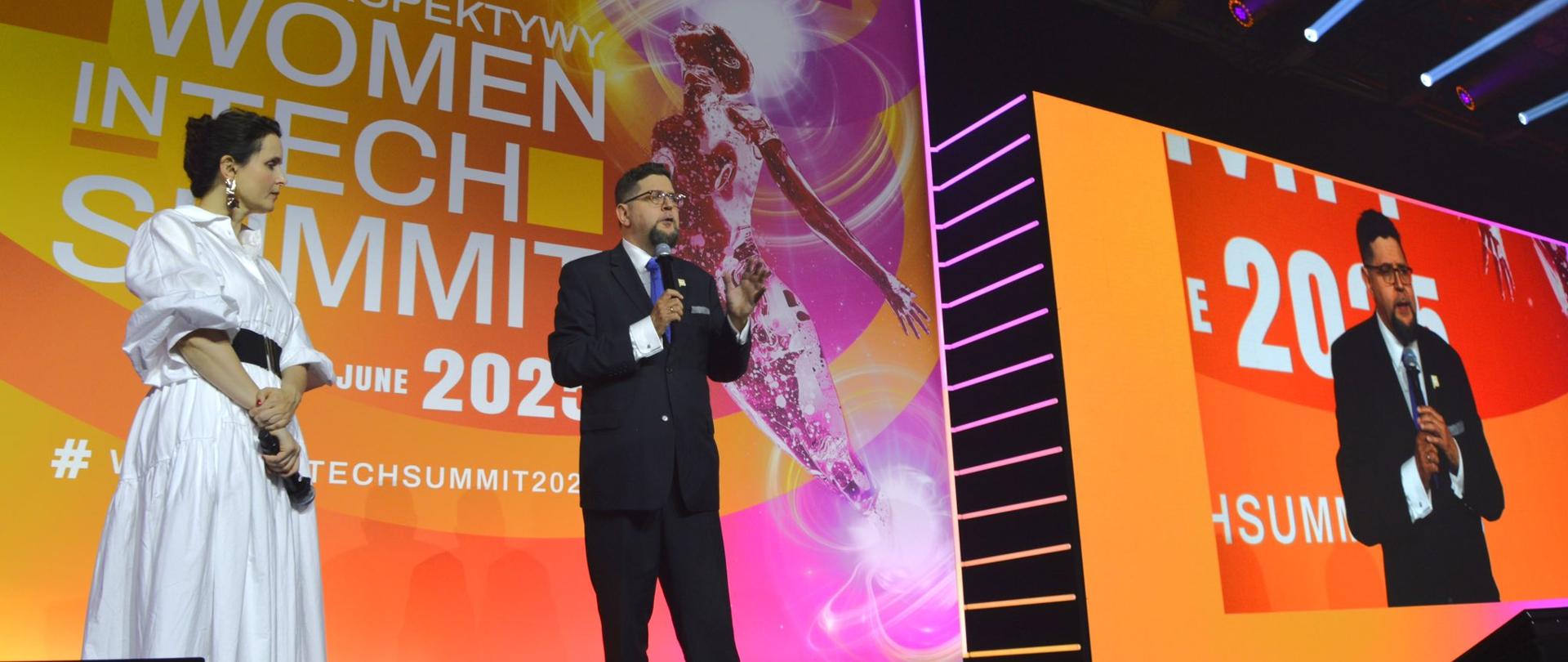 Za tle pomarańczowych ścian z napisem Women in Tech Summit 2025 stoi wiceminister Szeptycki i mówi do trzymanego w ręku mikrofonu, obok niego z lewej stoi kobieta w białej sukience.