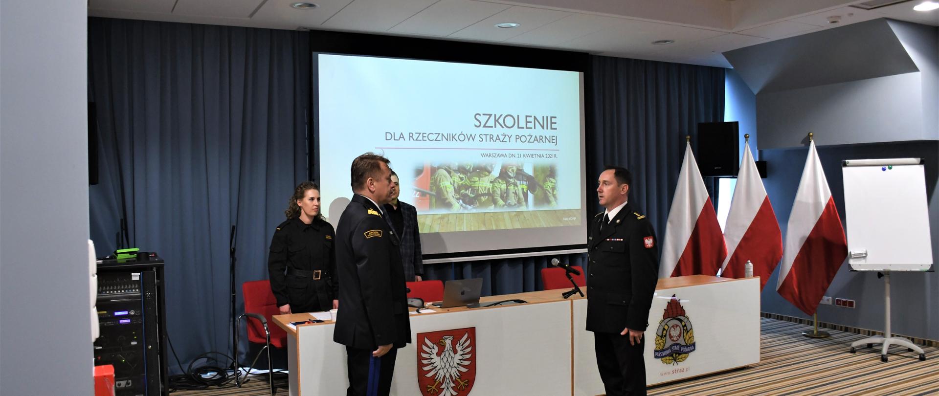 "Naczelnik Wydziału Organizacji i Nadzoru Komendy Wojewódzkiej PSP w Warszawie składa meldunek Mazowieckiemu Komendantowi Wojewódzkiemu PSP. Znajdują się na sali konferencyjnej. W tle wyświetlona jest prezentacja na ekranie.