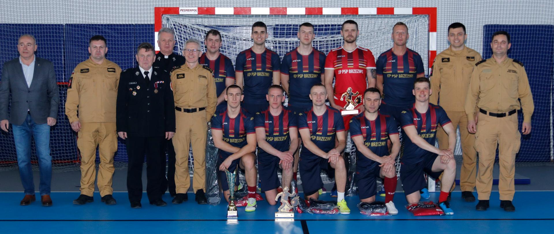 Reprezentacja komendy z kapelanem księdzem Władysławem Moczarskim, Burmistrzem Miasta Brzeziny Dariuszem Guzkiem, druhem Leszkiem Jarząbem Komendantem Powiatowym Państwowej Straży Pożarnej w Brzezinach starszym brygadierem Mariuszem Kaczmarskim i Zastępcą Komendanta Powiatowego Państwowej Straży Pożarnej w Brzezinach młodszym brygadierem Jakubem Brzózką pozują do wspólnego zdjęcia. Przed nimi na podłodze stoi zdobyty puchar za zajęcie pierwszego miejsca.