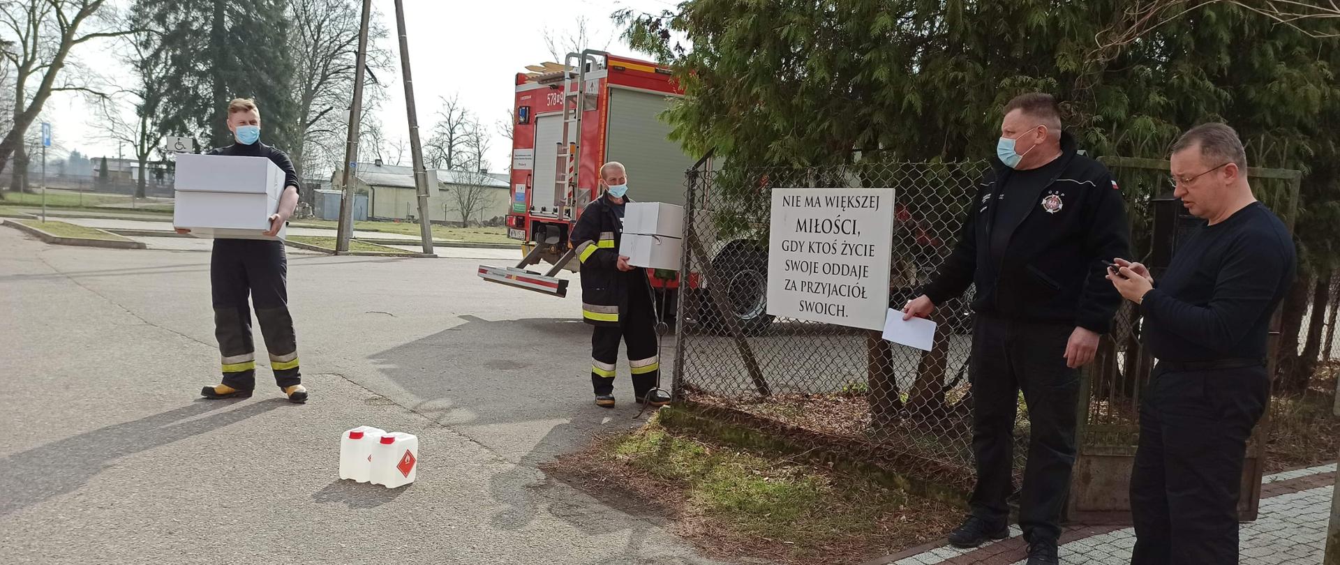 Z lewej strony dwóch strażaków niesie kartony z maseczkami, z prawej dwie osoby oczekujące na dostawę środków