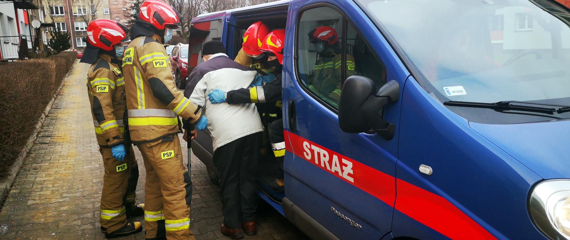 strażacy pomaga osobie starszej wsiąść do busa z napisem bocznym STRAŻ celem dowozu na szczepienie