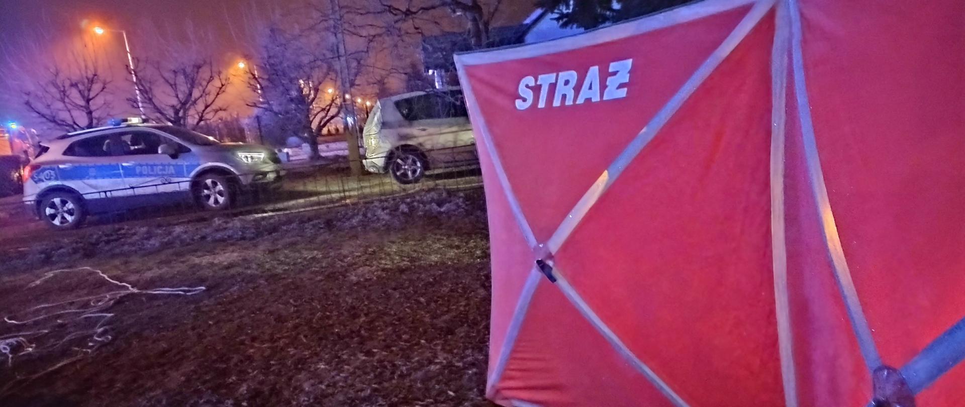 Zabezpieczony teren działań, strażacki parawan odgradzający miejsce akcji, w tle radiowóz policyjny. 