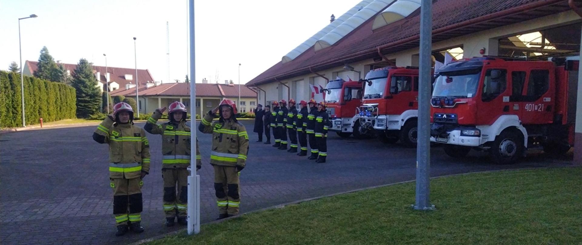 Na zdjęciu poczet flagowy składający się z trzech strażaków w ubraniu specjalnym, stojący przed masztem flagowym i salutujący.W tle pojazdy pożarnicze oraz pododdział strażaków oddających honor. 