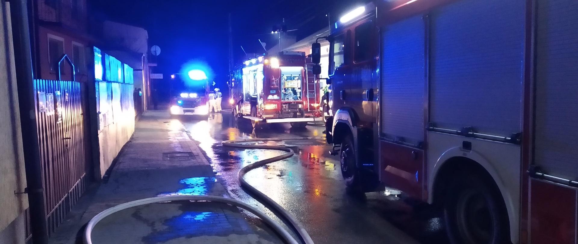 zdjęcie zrobione w nocy. Na ulicy stoją trzy samochody strażackie z włączonymi sygnałami świetlnymi na ziemi rozłożone węże strażackie napełnione wodą. 