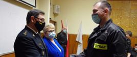 Widok z boku. Na pierwszym planie komendant powiatowy PSP w umundurowaniu podaje rękę druhowi OSP. Obok na drugim planie stoi wicestarosta wadowicka w niebieskim ubraniu cywilnym. W tle widoczni strażacy PSP i OSP w umundurowaniu. Wszystkie osoby w maseczkach ochronnych. 
