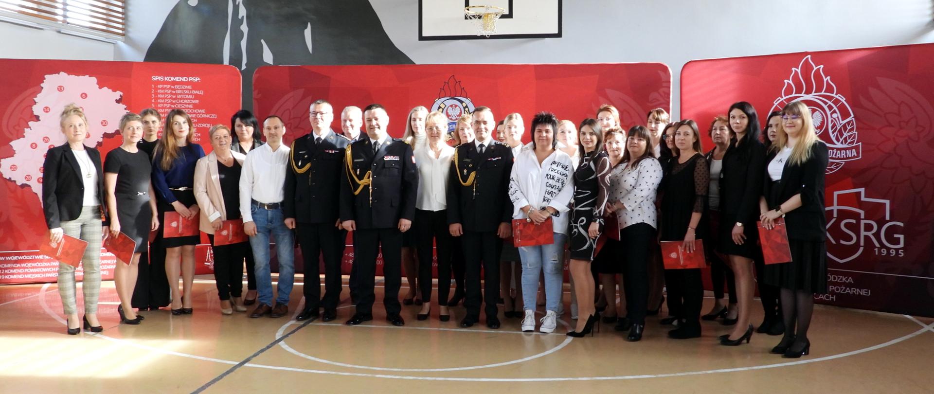 Na sali gimnastycznej, na tle czerwonej ścianki Komendy Wojewódzkiej, ubrani w mundury wyjściowe stoją w otoczeniu pracowników cywilnych: śląski komendant wojewódzki nadbryg. Jacek Kleszczewski wraz z dwoma zastępcami st. bryg. Mirosławem Synowcem i bryg. Arkadiuszem Krzemińskim. 