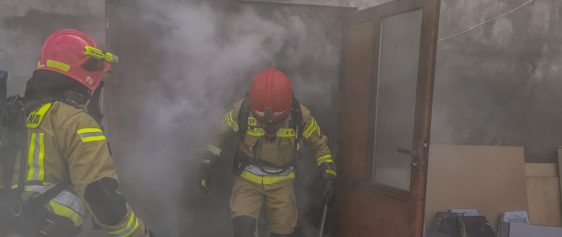 Pożar kotłowni na terenie zakładu produkcyjnego w Koźminie Wielkopolskim. Z otwartych drzwi wejściowych wydobywają się kłęby dymu. Strażacy w sprzęcie ochrony dróg oddechowych wychodzą z zadymionych pomieszczeń na zewnątrz. Przed obiektem stoi gaśnica wykorzystana do gaszenia pożaru oraz elementy wyniesione z kotłowni. 