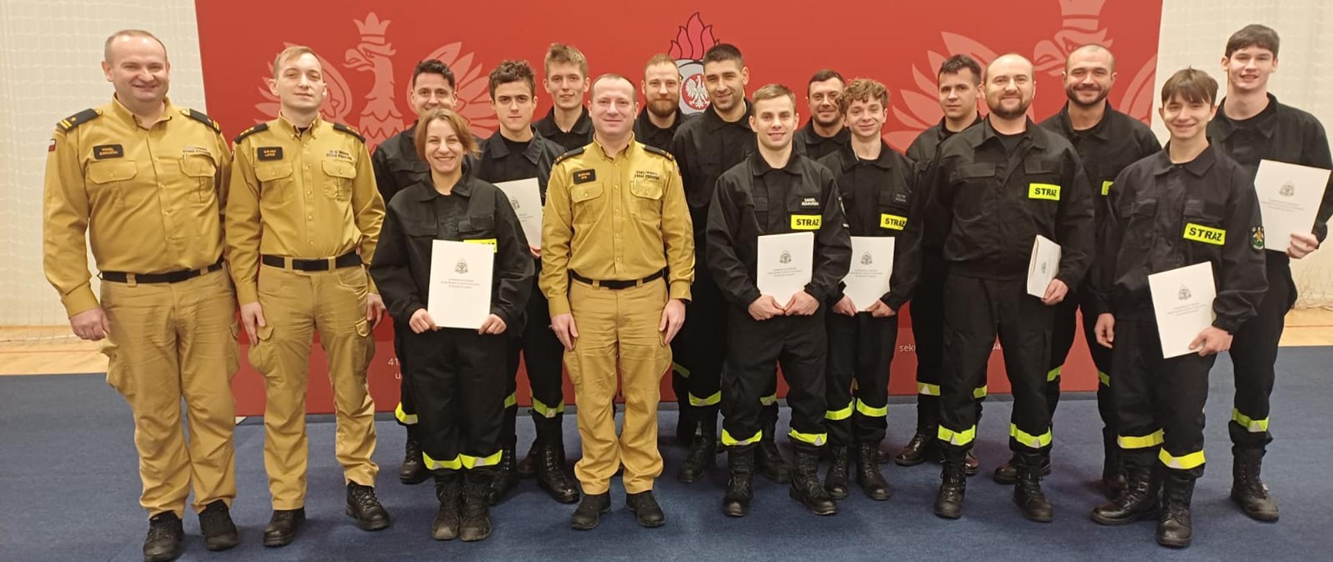 Grupa strażaków PSP i OSP w umundurowaniu stoi w sali gimnastycznej na tle czerwonej ścianki z godłem. Uczestnicy szkolenia trzymają w dłoniach zaświadczenia o ukończeniu kursu, pozując do wspólnego zdjęcia.