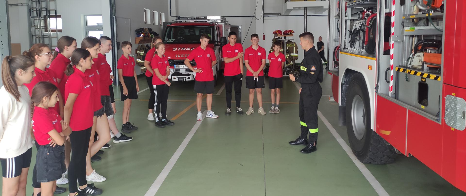 Wycieczka Młodzieżowej Drużyny Pożarniczej z OSP Nieciecz w KP PSP w Sokołowie Podlaskim - na zdjęciu młodzi adepci pożarnictwa zapoznają się z pojazdami i wyposażeniem znajdującym się w jednostce, przed czerwonym samochodem strażackim.