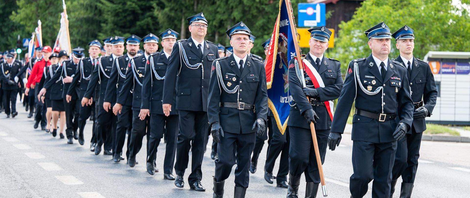 Zdjęcie przedstawia pododdziały OSP wraz z pocztami sztandarowymi w ugrupowaniu marszowym. Strażacy ubrani w mundur galowy OSP.