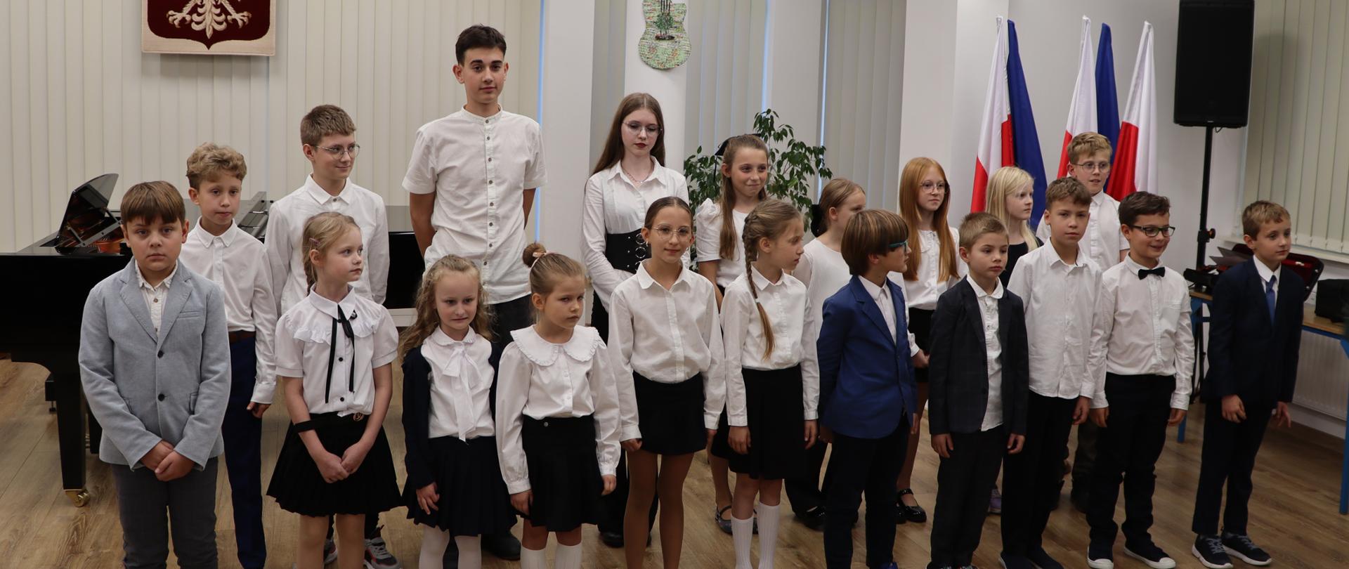 W sali, ustawione w dwóch rzędach, stoją dziewczynki i chłopcy. Dzieci ubrane są w białe koszule oraz ciemne spodnie lub spódnice. Czworo chłopców ma założone marynarki. Za nimi czarny fortepian oraz wiszące na ścianie godło Polski. Biały orzeł na czerwonym tle.