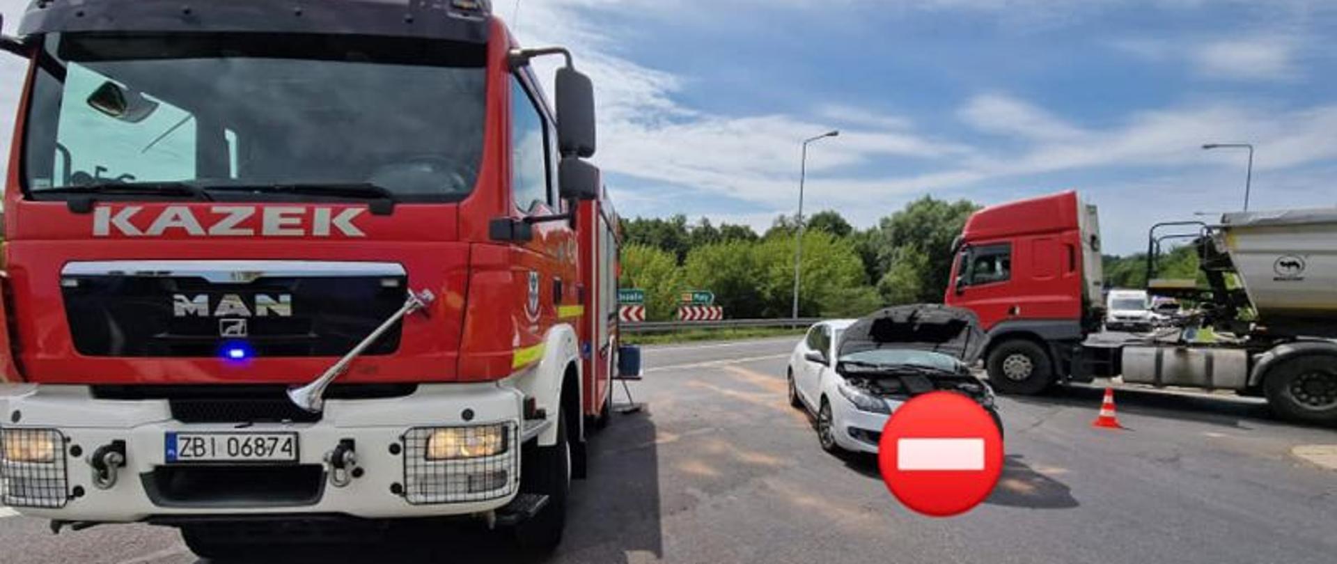 Zdjęcie wykonane w dzień. Centralny punkt zdjęcia stanowi rozbite białe auto. Po lewej widoczny wóz strażacki w tle ciężarówka stojąca na skrzyżowaniu.