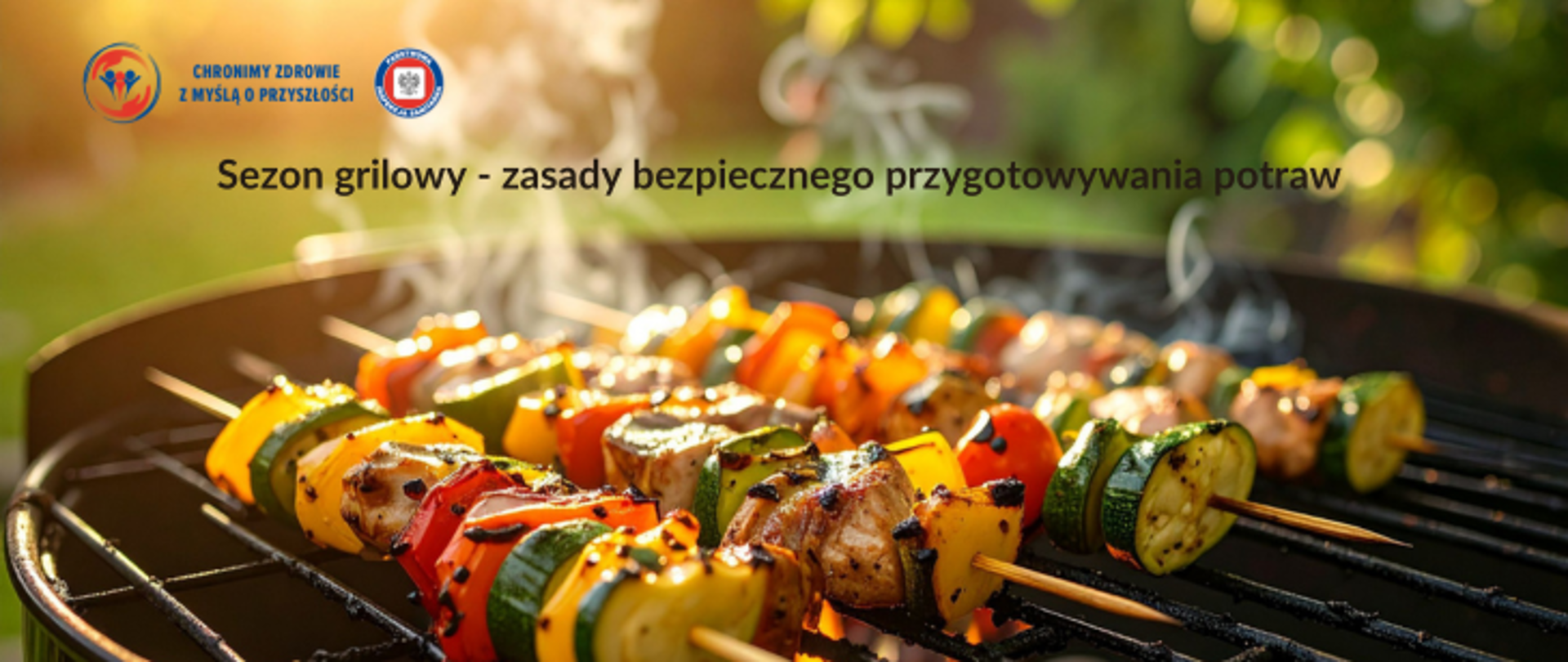 Na zielono-brązowym tle na pierwszym planie grill. Na nim cztery szaszłyki warzywno-mięsne. Powyżej ciemny napis: Sezon grillowy - zasady bezpiecznego przygotowywania potraw. W lewym górnym rogu logo Państwowej Inspekcji Sanitarnej wraz z hasłem "Chronimy zdrowie z myślą o przyszłości. 