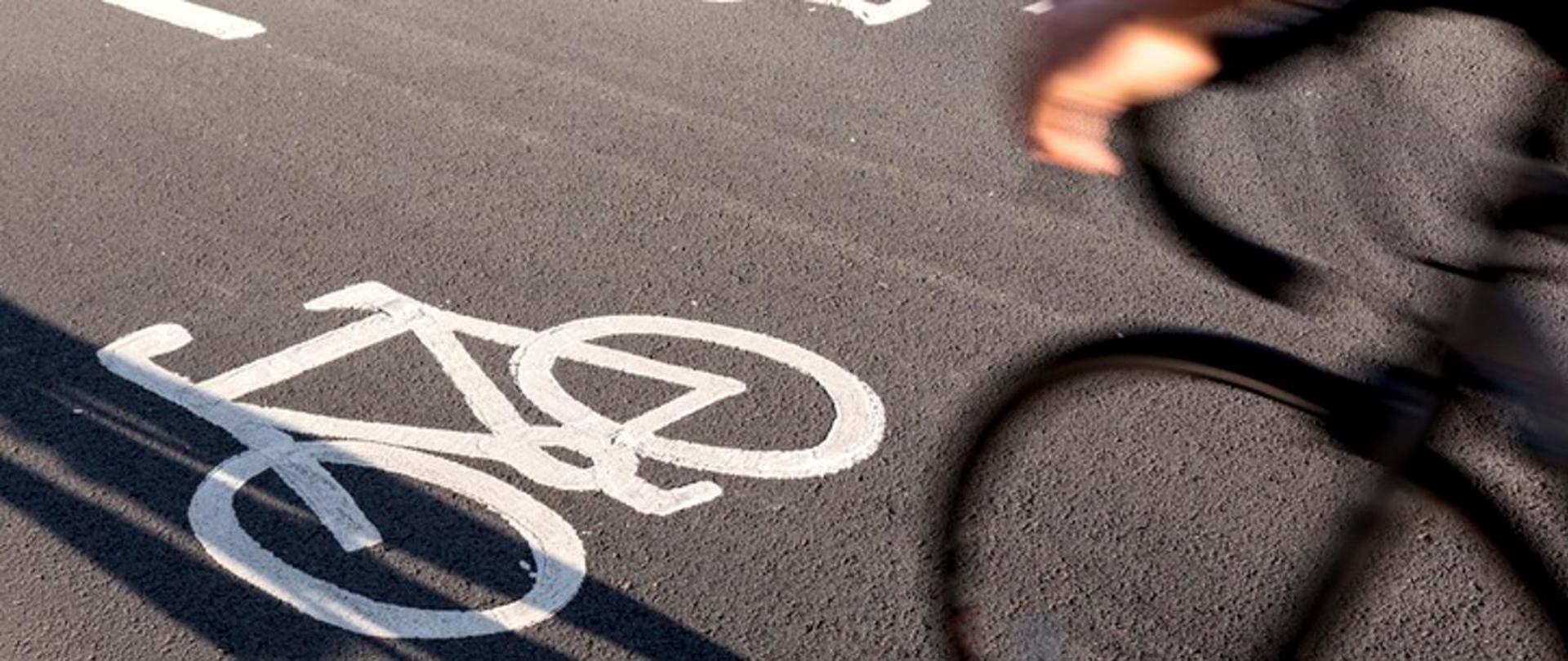 Osoba dojeżdżająca do pracy na ścieżce rowerowej. Na czarnej jezdni narysowany biały rower. Na pierwszym widok koła rowerowego i rąk na kierownicy.