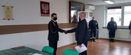 W tle ściana świetlicy z wiszącym na niej polskim godłem narodowym oraz rotą ślubowania. Dalej na prawo stoją strażacy zmiany służbowej. W centrum zdjęcia Komendant Powiatowy PSP gratuluje nowemu strażakowi przyjęcia w szeregi PSP Strażacy mają na twarzach maseczki. 