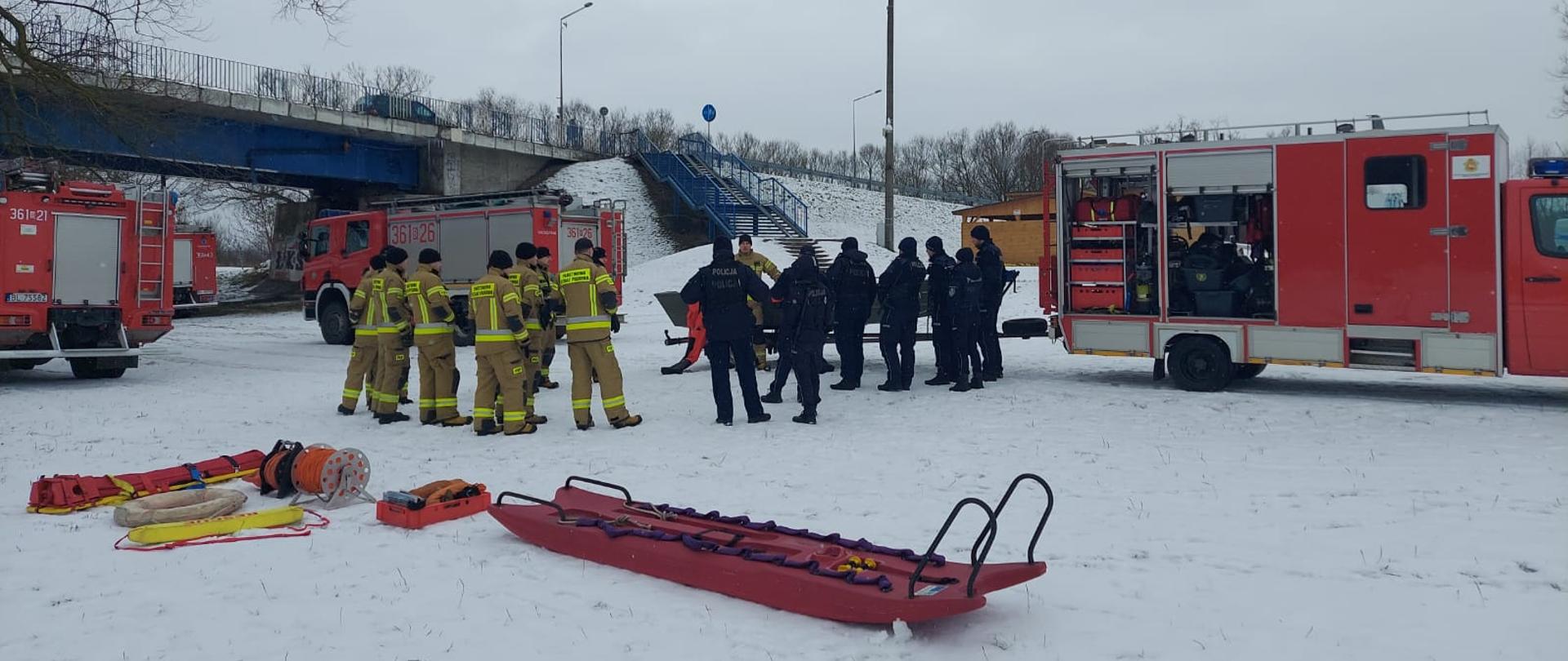 Strażacy razem z policją omawiają zakres ćwiczeń na lodzie. W tle widok mostu oraz samochody pożarnicze.