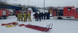 Strażacy razem z policją omawiają zakres ćwiczeń na lodzie. W tle widok mostu oraz samochody pożarnicze.